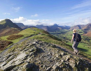 Woman hiking on a rocky hilltop path overlooking rolling green valleys and distant peaks in the Lake District on a clear, sunny day. ?? ''
