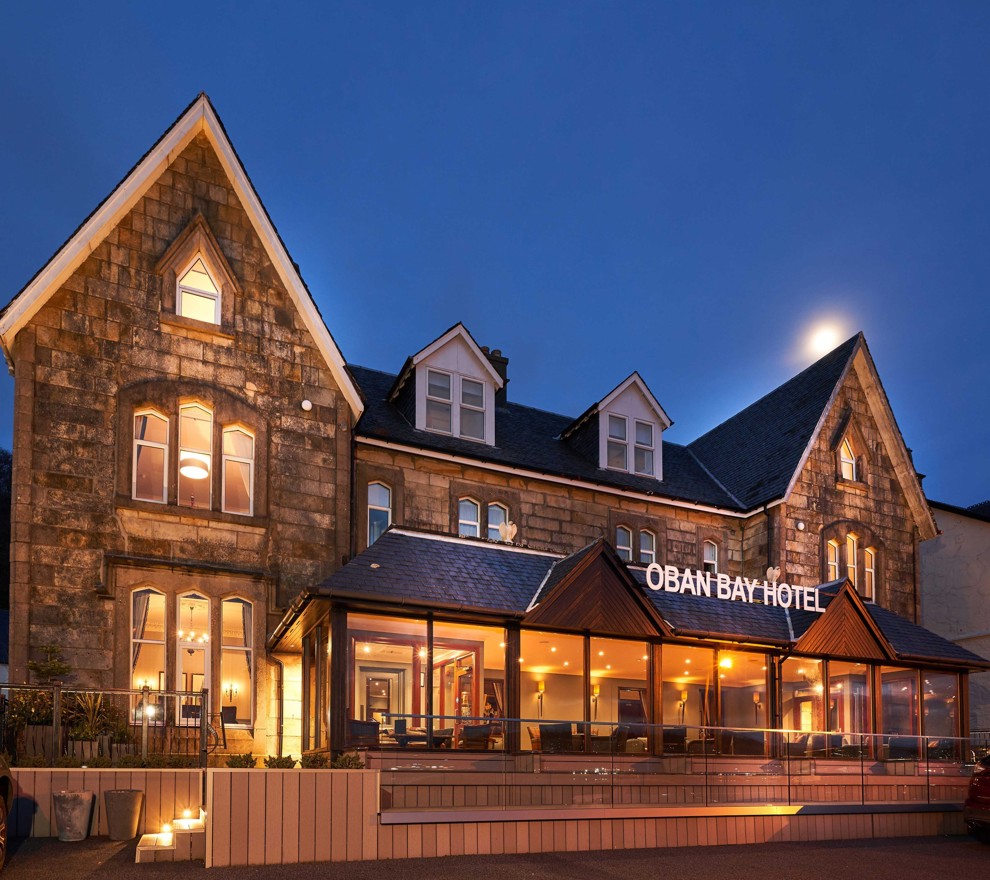 Exterior view of the Oban Bay Hotel, a traditional stone building with a modern glass extension, illuminated at dusk with a clear night sky. ?? ''